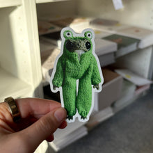 Load image into Gallery viewer, A hand holds a die-cut sticker of a green frog wearing a onesie against a blurred background of white shelves and boxes.