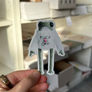 A hand holds a die-cut sticker of a green frog wearing a white t-shirt with the text 'FUCK OFF' in a real storage room with shelves and boxes in the background.