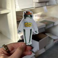A hand holds a small die-cut sticker or card featuring a cartoon frog character with a yellow shirt and orange hat, in front of white shelving filled with boxes and papers.